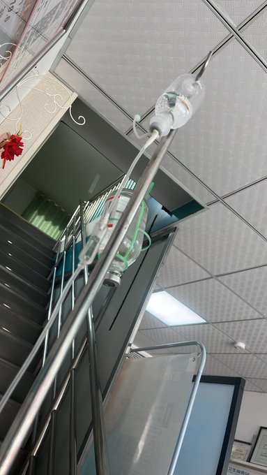 A photo taken at a low angle showing an IV drip stand with fluid bags hanging, set against a hospital ceiling and metal railing.