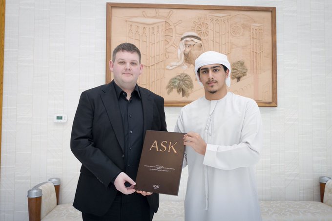 The image features two men standing next to each other with one man holding a book titled "ask" while the other man holds a sign that says "ask". The background includes a painting of a mosque, which suggests that they are in an Arabic-speaking country or region. The text on the post indicates that there is a joint venture between Keeta and ASK Group, led by Sheikh Ahmed bin Sultan bin Khalifa bin Zayed Al Nahyan, aiming to tokenize tens of billions of dollars of commodities and modernize cross-border transactions.