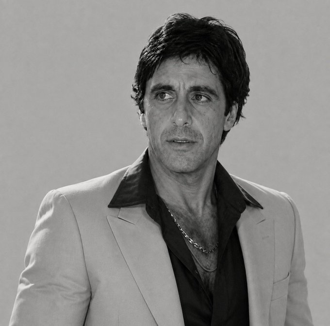 A black-and-white portrait photograph of a man with dark, wavy hair wearing a light-colored blazer over a dark, open-collared shirt and a silver chain necklace.