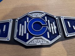 A close-up photograph of a blue and silver championship wrestling belt plate featuring the letter 'C' in the center.
