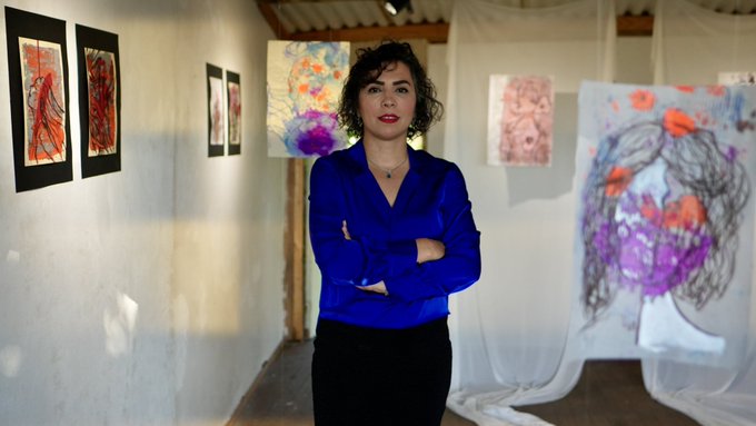 A woman in a blue blouse stands with arms crossed in an art gallery, surrounded by colorful abstract paintings displayed on walls and hanging fabric.