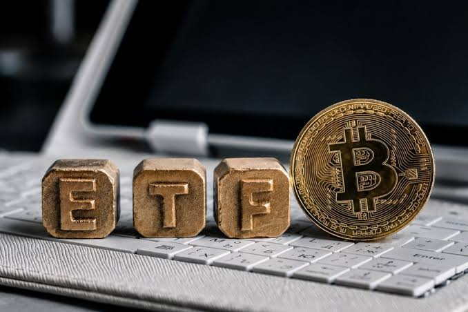 A physical Bitcoin token stands next to three wooden blocks spelling 'ETF' on a laptop keyboard.