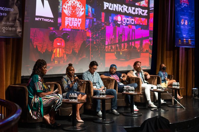 A panel of six people sits on a stage at a conference, with a large screen behind them displaying logos and artwork including 'MYTHICAL,' 'DREAM FURY,' 'PUNKRAIZERZ,' and 'gaia.'
