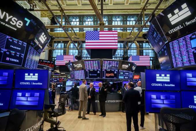 The image shows a group of people standing around a desk inside a building, possibly a stock exchange or financial institution. They are surrounded by multiple screens displaying various stock market information, including the Dow Jones Industrial Average (DJIA) and other stock tickers. The scene suggests that these individuals might be discussing or analyzing the stock market data displayed on the screens.

The post about U.S. stock trading and tokenized shares for users outside the United States implies that this gathering could be taking place in a location where such trading is allowed, possibly within the United States. It also indicates that the focus of their discussion might be on the potential benefits or risks associated with these new types of financial instruments.