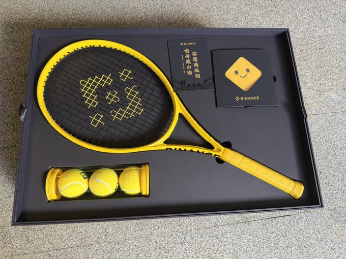 A yellow and black Binance-branded tennis racket set with tennis balls and accessories displayed in a black box.