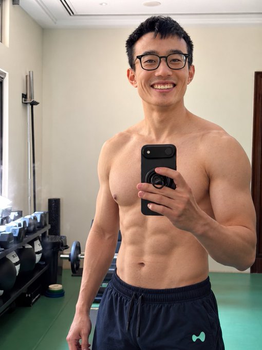 A shirtless, smiling man with glasses takes a mirror selfie in what appears to be a gym, showing his fit physique.