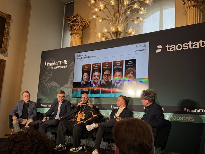 The image shows a group of five people sitting around a table, engaged in conversation. They are all wearing suits and ties, suggesting that they might be attending an important event or meeting. The backdrop features a large screen displaying a list of names on it, which could be related to their discussion. The room has a grand appearance with columns and a chandelier hanging from the ceiling, giving it an elegant atmosphere.