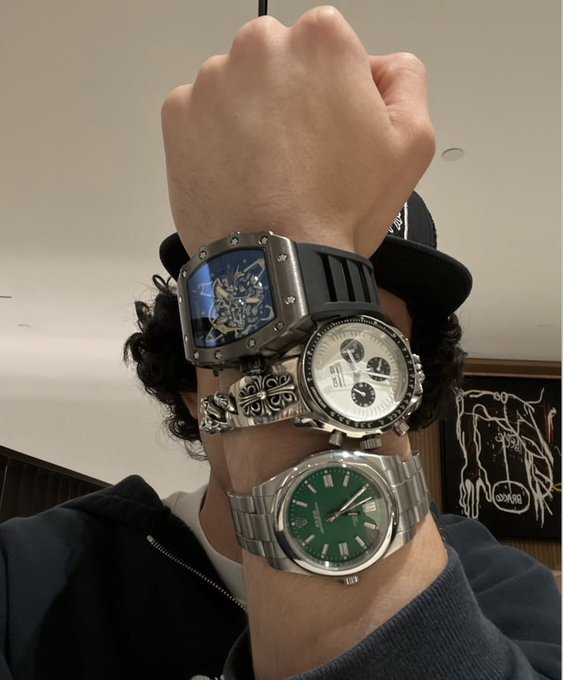 A person shows off their wrist stacked with three luxury watches: a Richard Mille, a chronograph, and a Rolex Oyster Perpetual with a green dial.