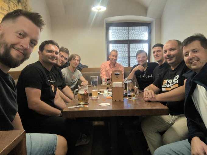 A group of eight smiling men sit around a wooden table in a restaurant or pub setting, with beer glasses and a menu visible.