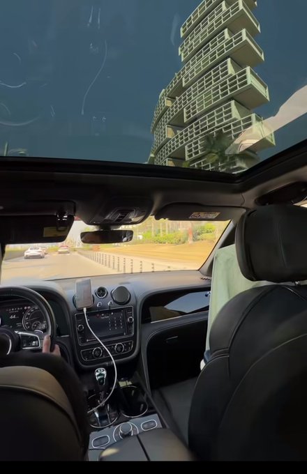 Interior view from the backseat of a luxury car with a glass roof, showing the driver’s hands on the steering wheel, the dashboard with a mounted phone, and a tall modern high-rise building visible outside the windshield, perfectly matching the post’s vibe of a focused “GM” (good morning) drive to start the day.