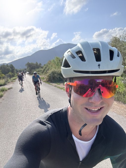 A selfie of a man wearing a white cycling helmet and red KOO brand sunglasses, with other cyclists riding on a road behind him and mountains in the background.