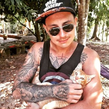 A tattooed man wearing sunglasses, a 'Space Invaders' cap, and a black tank top holding an orange cat outdoors.