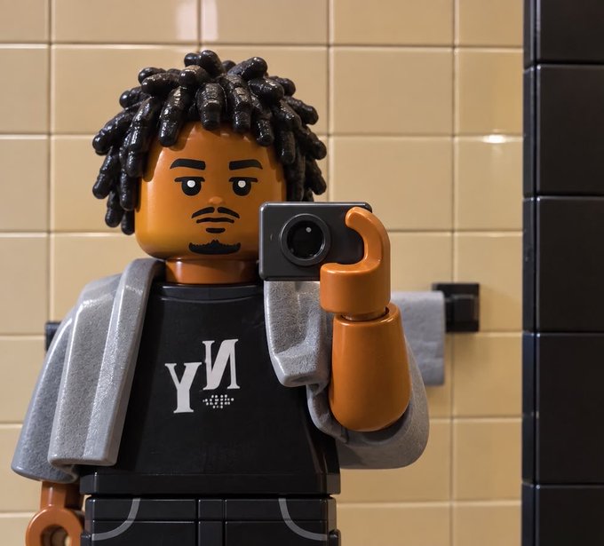 A close-up of a Lego-style minifigure with brown skin, black dreadlocks, and a beard, wearing a black shirt with a white 'M' logo and a gray jacket. The figure is holding a black camera, set against a background of beige subway tiles.