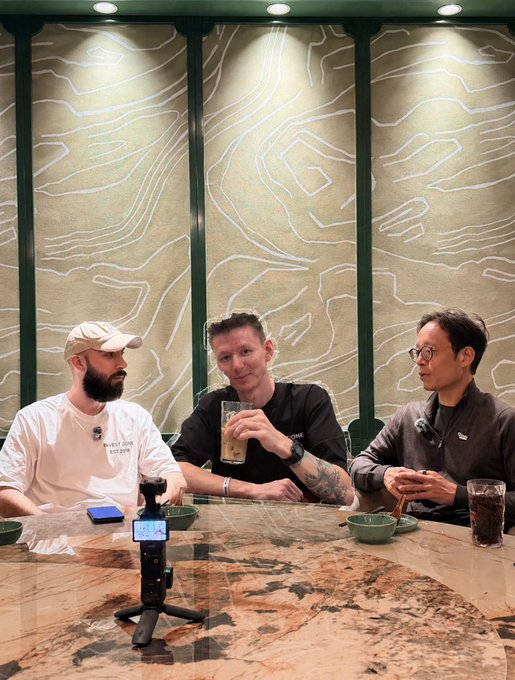 Three men sitting around a marble table in what appears to be a podcast or video recording setup, with one man in a 'West Zone Studios' shirt and beige cap, another in a black shirt holding a drink, and a third in a dark jacket.