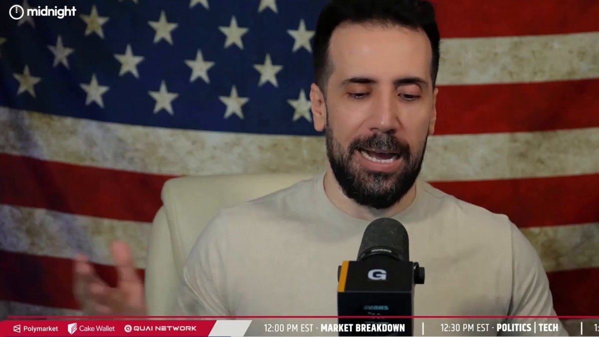 The image features a man wearing a white shirt sitting behind a microphone at a news broadcast station. He appears to be talking about something related to MicroStrategy's latest $BTC movement or Bitcoin. The background of the image includes an American flag, which adds context to the setting and suggests that this might be a news broadcast on TV.