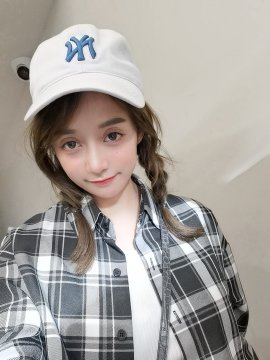 A young woman takes a selfie wearing a white NY (New York Yankees) cap and a plaid shirt, with her hair in loose braids. The post playfully compares her 2022 and 2026 appearances, inviting followers to spot the differences in her look over the years as part of the #Bitget看球狂欢季 (Bitget Watching the Game Festival).