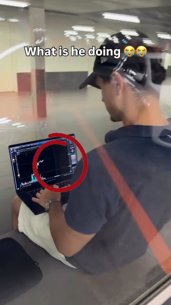 A man wearing a cap sits in what appears to be a parking garage using a laptop displaying trading charts, with overlaid text reading 'What is he doing' with crying emojis and a red circle drawn on the screen.