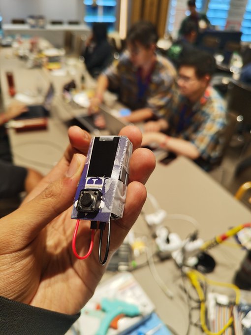 A hand holds a small blue and white electronic device labeled 'UNITREE Go1' in what appears to be a hackathon or workshop setting with people working at tables in the background.