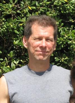 The image features a man wearing a gray shirt with his face turned towards the camera. The background of the photo shows green bushes, suggesting that they are outdoors or possibly in an area with some greenery. There is also text present in the image, which reads "History: 27 years ago, Bitcoin pioneer Hal Finney presented SHA-1 zero-knowledge proof at Crypto Conference." This information provides context to the man's appearance and the setting of the photo.