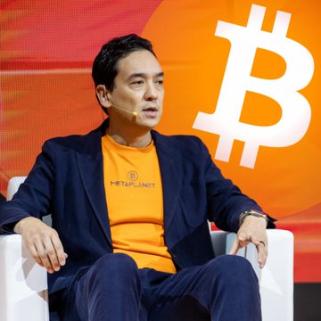 A photograph of a man wearing a yellow 'METAPLANET' t-shirt and dark blazer, sitting on a white chair in front of a bright orange backdrop featuring a large white Bitcoin logo.