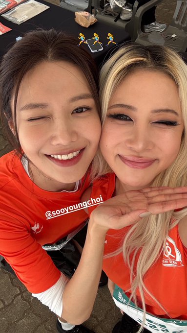 Two smiling women taking a selfie, both wearing red shirts with running bibs visible; one shirt displays '@sooyoungchoi' and the bib shows 'SK', with small running figure emojis on the table behind them.