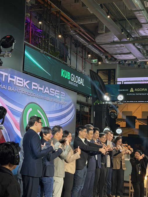 A group of men in suits pose with thumbs up at a corporate event, with 'THBK PHASE 2' and 'KUB GLOBAL BANGKOK' signage visible in the background.