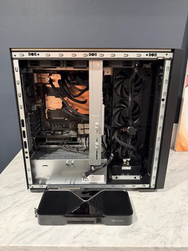 An open computer case sits on a white surface displaying internal components including cooling fans, cables, and a mounted GPU, with a small black device in the foreground.