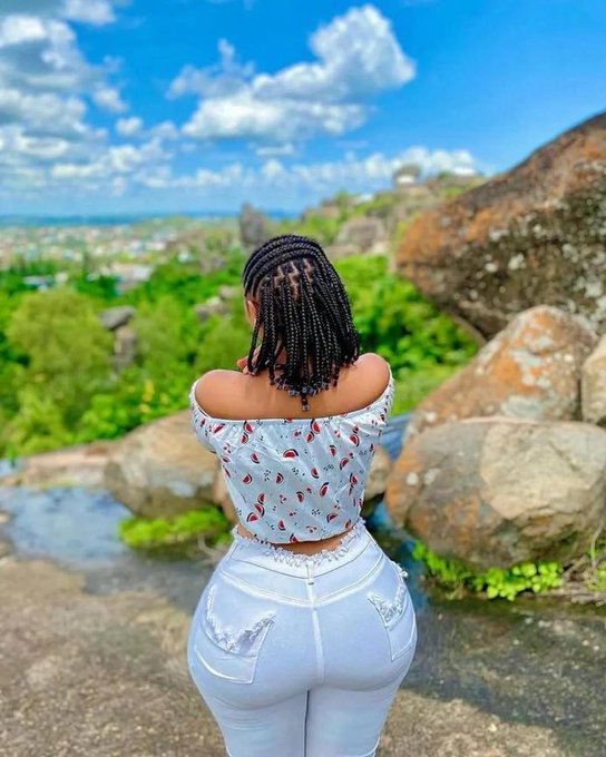 A woman with long braided hair wearing an off-shoulder patterned crop top and white jeans stands with her back to the camera, overlooking a scenic landscape of green hills and large boulders.