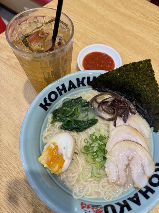 A bowl of ramen at KOHAKU restaurant featuring chashu pork, a soft-boiled egg, spinach, mushrooms, and nori, served with a side of red sauce and iced tea.