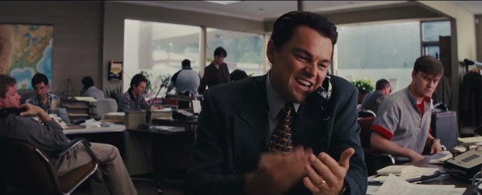 A scene from a movie showing Leonardo DiCaprio in a suit talking on a phone and laughing in a busy office environment, with other workers at desks in the background.