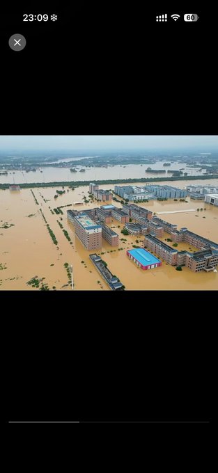 An aerial photograph showing a severely flooded area with brown water submerging roads, farmland, and surrounding multiple multi-story buildings under an overcast sky, displayed as a phone screenshot at 23:09.