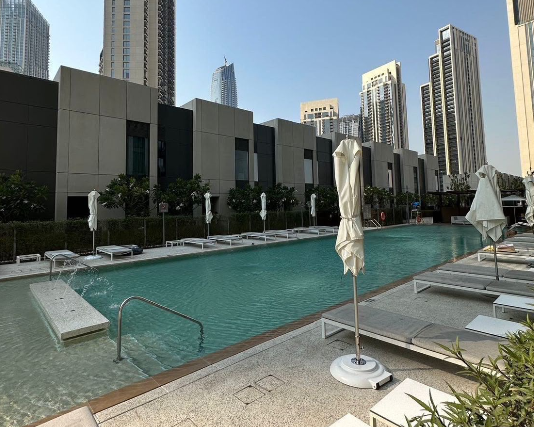 An outdoor swimming pool at a modern residential complex is surrounded by lounge chairs and closed white umbrellas, with high-rise city buildings visible in the background.