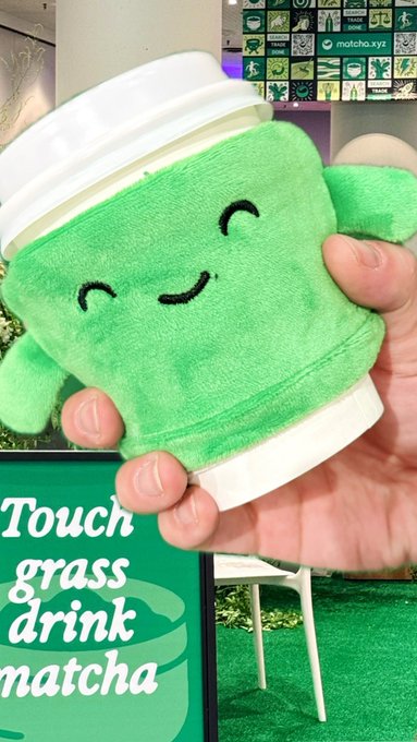 The image features a person holding up a green cup with a smiley face design, which appears to be made of plush material or stuffed animal-like fabric. The background shows a green grass field, suggesting that this scene takes place outdoors. There is also an advertisement for "catch y'all tomorrow @ethconf", indicating the image was taken at an outdoor event or gathering related to ethical consumerism and sustainability.