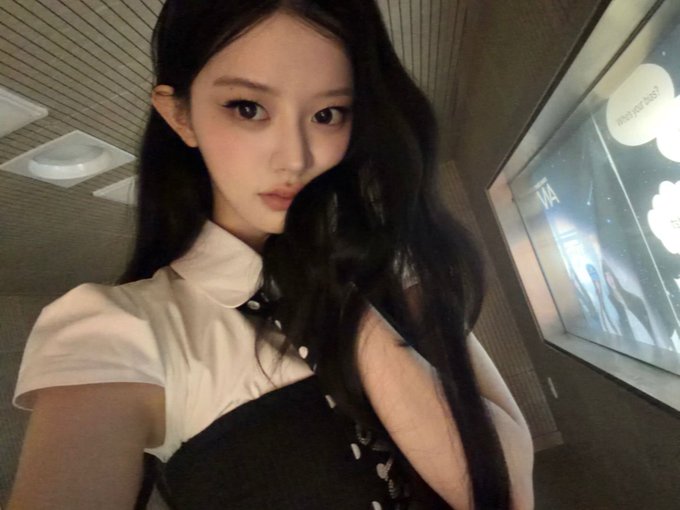 A selfie of a young woman with long dark hair wearing a white and black outfit, posing in what appears to be a hallway.