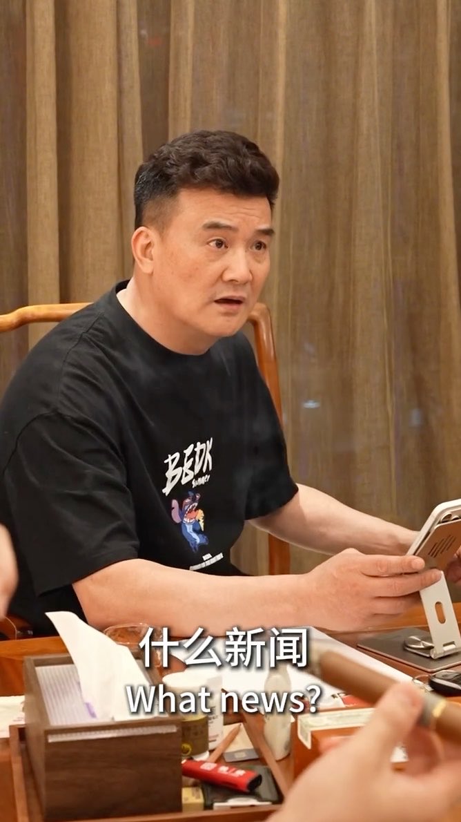 A man in a black graphic t-shirt sits at a desk holding a tablet, with subtitles reading '什么新闻 / What news?' on the screen.