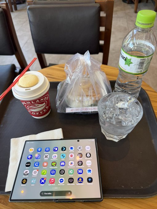 A tray on a cafe table holding a Highlands Coffee cup, a plastic bag with food, a glass of ice water, a bottled water, and a Samsung tablet displaying a screen full of app icons.
