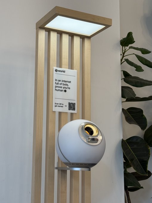 A spherical white device mounted on a wooden stand beneath a square light panel, featuring a 'world' poster with a QR code reading 'In an internet full of bots, prove you're human. Scan QR to get started.'