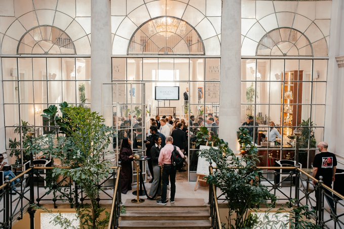 An interior view of an elegant venue with large arched windows, where a crowd of people is gathered, possibly at an event or boutique space with plants and clothing displays.
