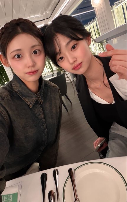 A selfie of two young women posing closely together at a restaurant table set with white plates, black-handled cutlery, and chopsticks.