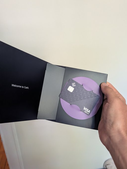 A hand holding open a dark envelope/package that reads "Welcome to Cash." on the left side, revealing a dark "Cash App" Visa debit card nestled in a purple circle on the right.