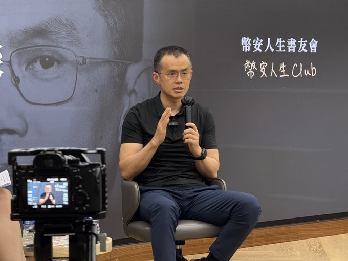 A man with glasses in a black polo shirt speaks into a microphone while seated, with Chinese text '幣安人生書友會' (Binance Life Book Club) on the wall behind him.