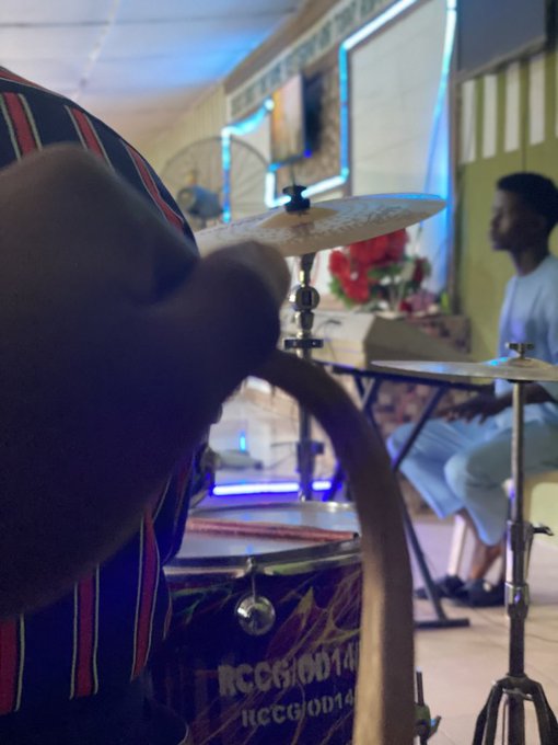 A person plays drums with a cymbal labeled 'RCGGOD14' visible, while another person sits at a keyboard in the background of what appears to be a church or performance space.