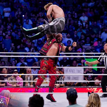 A muscular professional wrestler in red and gold pants lifts a shirtless opponent high into the air in a stacked lifting slam, while another wrestler and a referee watch from the ring. A fan sign reading "WE ARE LOVE AND IGNITE" is visible in the crowd behind the ropes. The image captures the power and spectacle that aligns with the post's emphasis on the wrestler's imposing size and strength.