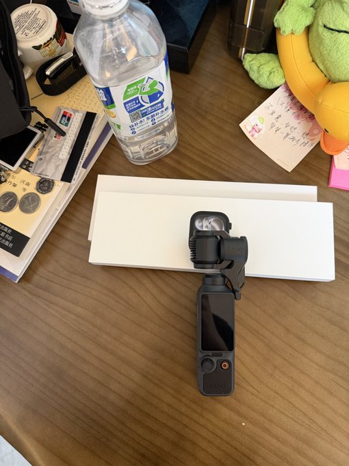 A DJI Osmo Pocket gimbal camera sits on a white box on a wooden desk, next to a clear plastic water bottle, papers, and a green plush toy.