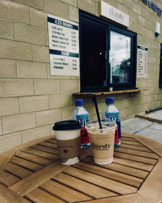 Two iced drinks from "thirsty" cafe (a coffee and a cooler) sit on a wooden table with water bottles, ready for a morning crypto trading session. The menu behind shows iced drinks at $3.95 and breakfast items like acai bowls and porridge for $8.95—fuel before diving into the charts.