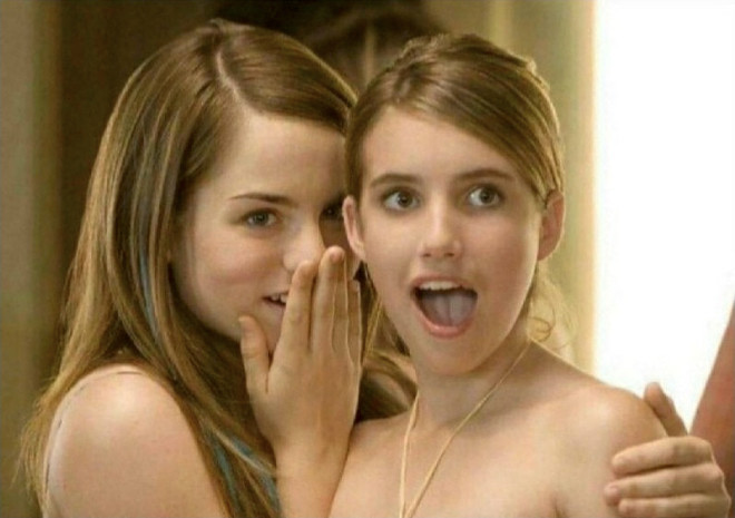 Two young women with long hair are shown closely together, with one whispering into the other's ear while the second woman reacts with an exaggerated expression of surprise.