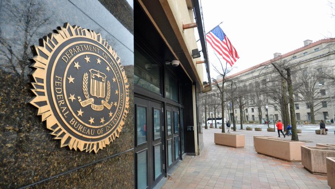 An outdoor view shows the seal of the Department of Justice Federal Bureau of Investigation on a building wall, with an American flag hanging nearby.