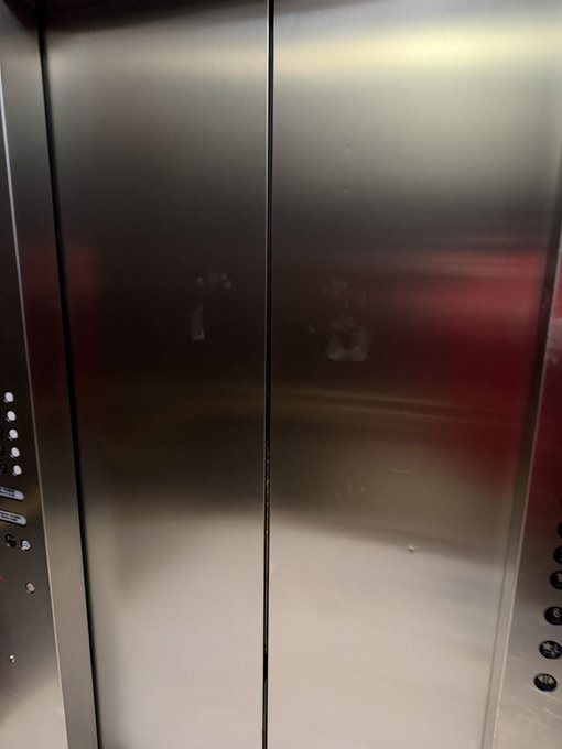 A view of closed, brushed metal elevator doors reflecting a reddish light, with elevator control buttons visible on the right side panel.