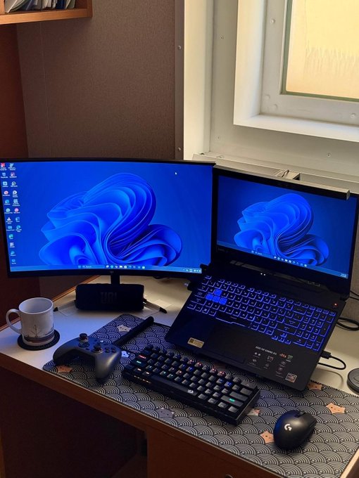 A gaming/workstation setup with a curved monitor and laptop both displaying the Windows 11 blue bloom wallpaper, accompanied by an Xbox controller, mechanical keyboard, and wireless mouse—suggesting the "GM CT (Good Morning Central Time)" poster is starting another day at their dual-screen desk.