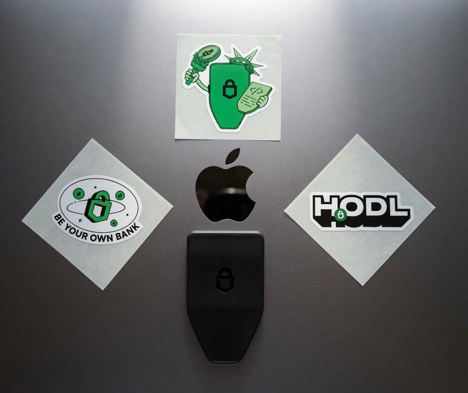 The image features a collection of stickers with various designs, including one that has a Statue of Liberty vibe. The stickers are attached to an Apple computer using a black keyring or lock. The stickers include "Be your own bank" and "HODL". One of the stickers is a keychain shaped like a Statue of Liberty.